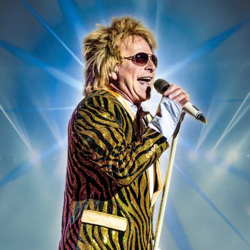 A male singer wearing a gold-and-black sequined jacket and sunglasses performs on stage, holding a microphone and singing energetically against a backdrop of bright blue lights.