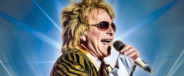 A male singer wearing a gold-and-black sequined jacket and sunglasses performs on stage, holding a microphone and singing energetically against a backdrop of bright blue lights.