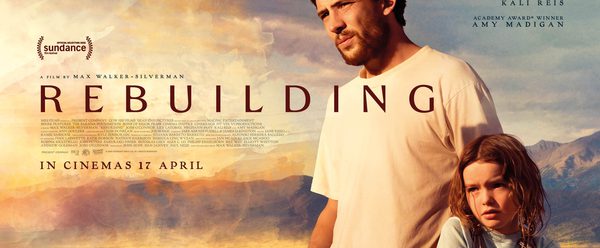 a man and a young girl standing together in a sunlit rural landscape with mountains in the background. The man, with short dark hair and light facial hair, looks off into the distance with a serious expression, while the girl stands close beside him, appearing thoughtful.