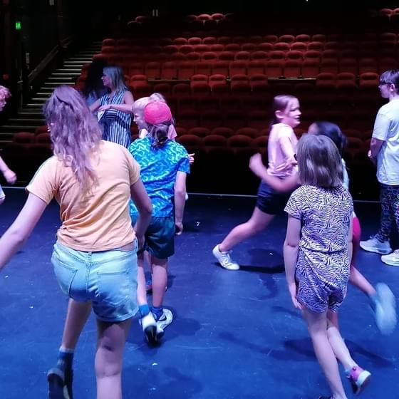 A group of young people dance around on a stage, facing out into the theatre's auditorium