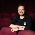 Mat sitting in between 2 rows with his arms folded over the seats in front of him, smiling at the camera.