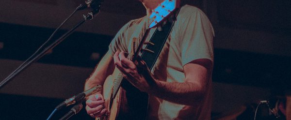 A musician performs on stage, holding an acoustic guitar, under soft lighting with decorative bunting in the background.