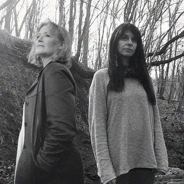 Two figures stand side by side in a wooded area, surrounded by bare tree branches, captured in black and white.