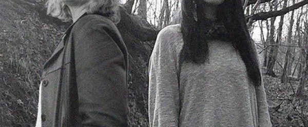 Two figures stand side by side in a wooded area, surrounded by bare tree branches, captured in black and white.