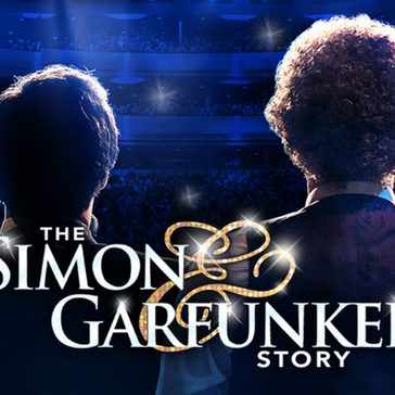 Two musicians seen from behind stand on a brightly lit stage facing a large audience in a theater, with the title “The Simon & Garfunkel Story” displayed across the image.