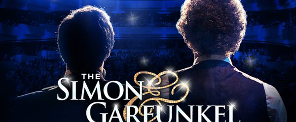 Two musicians seen from behind stand on a brightly lit stage facing a large audience in a theater, with the title “The Simon & Garfunkel Story” displayed across the image.