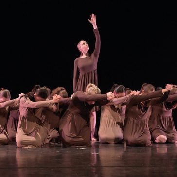 Dancers holding move with one standing in the middle holding arm up and the rest surrounding her kneeling.