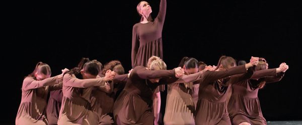 Dancers holding move with one standing in the middle holding arm up and the rest surrounding her kneeling.