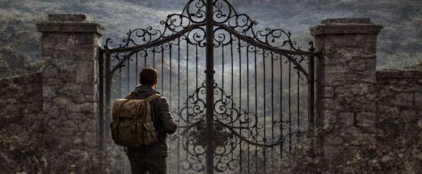 A lone traveler with a backpack stands before an ornate iron gate, looking toward a large, mist-shrouded mansion nestled in a distant valley under a cloudy sky.