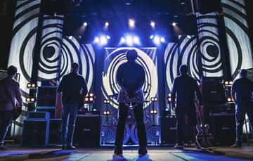 Five male musicians stand on stage with their backs to the audience, facing a large wall of amplifiers and psychedelic black-and-white swirl backdrops. The central figure stands with hands behind his back, while others hold guitars. Blue and white stage lights beam down from above.