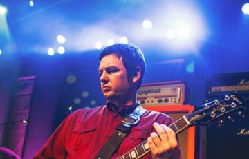 Person in a red shirt plays an electric guitar on stage, with amplifiers in the background and colourful stage lighting above.