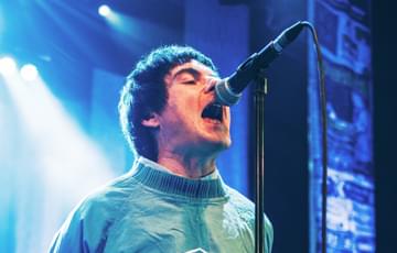 A male singer wearing a teal Umbro jacket performs energetically with his mouth open close to a microphone. His hands are behind his back. In the background, another man plays an acoustic guitar under blue stage lighting.