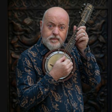 Bill holding a small banjo-like instrument, looking directly at the camera, wearing a patterned dark shirt against an ornate carved background.