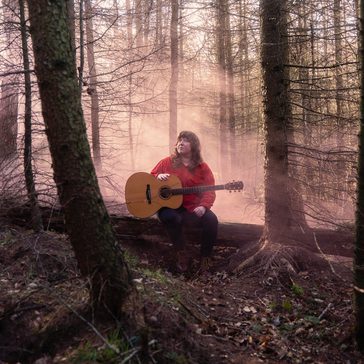 Holly sat on a log in a serene forest, playing an acoustic guitar amidst soft, misty light filtering through the trees.