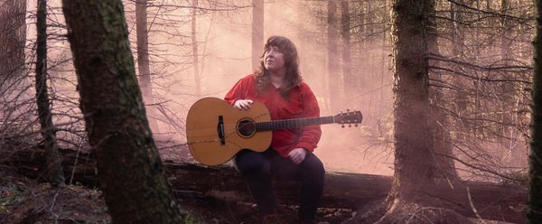Holly sat on a log in a serene forest, playing an acoustic guitar amidst soft, misty light filtering through the trees.