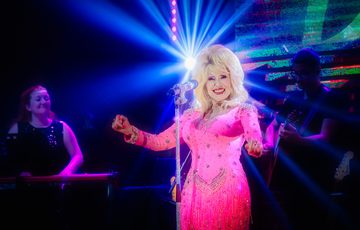 A glamorous female singer in a sparkling pink dress performs on stage, smiling at the audience under bright blue and purple lights, with band members playing instruments in the background.