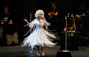 A female performer with big curly blonde hair sings into a microphone on stage, wearing a white fringed dress and high heels, with a live band playing in the background under stage lights.