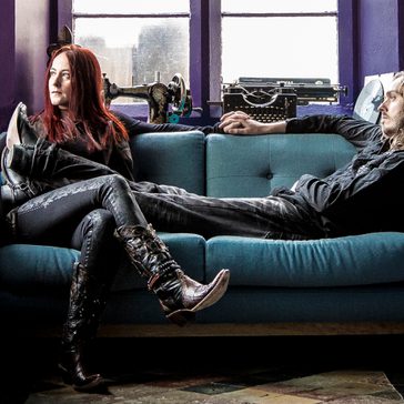 A man and woman sit on a teal sofa in a purple room with a window, sewing machine, and typewriter; the woman wears black clothing and elaborate cowboy boots, and the man wears black clothing, with long hair.