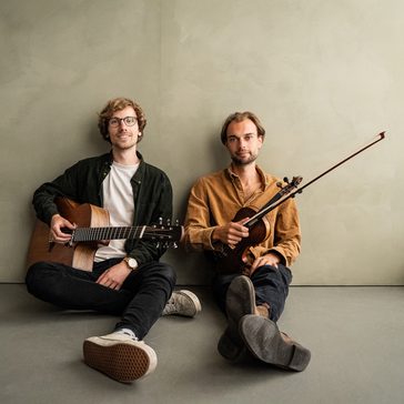 Two musicians seated on the floor against a minimalist wall, one with an acoustic guitar and the other with a violin, posing with their instruments.