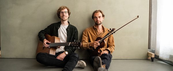 Two musicians seated on the floor against a minimalist wall, one with an acoustic guitar and the other with a violin, posing with their instruments.