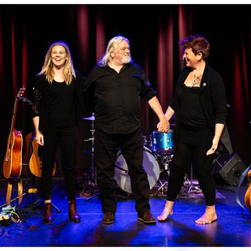 Three performers dressed in black stand on stage holding hands and smiling, with guitars, a drum kit and red curtains behind them.