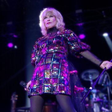 Singer Toyah performing on stage in a sparkling multicolored dress under purple concert lights while holding a microphone stand.