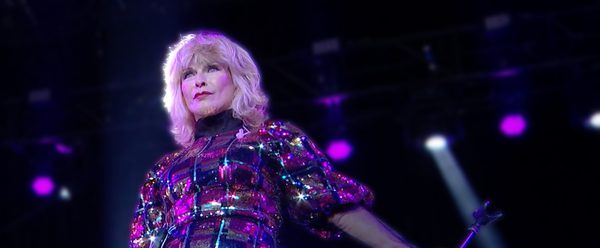 Singer Toyah performing on stage in a sparkling multicolored dress under purple concert lights while holding a microphone stand.