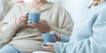 Two women sit chatting, each holding a cup of coffee, on a comfortable sofa