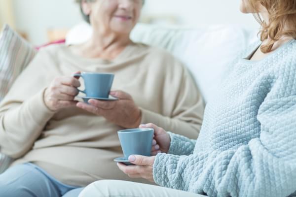 Two women sit chatting, each holding a cup of coffee, on a comfortable sofa